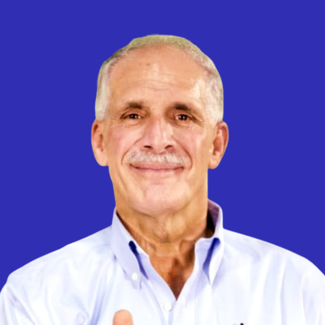 Un hombre mayor con pelo corto y canoso y bigote sonríe. Lleva una camisa de cuello azul claro y posa delante de un fondo azul sólido.