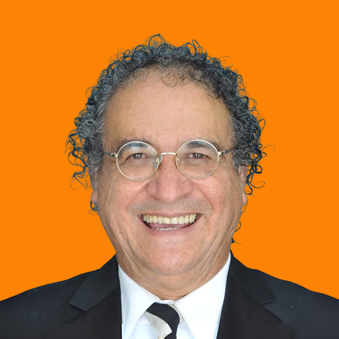 Un hombre sonriente con el pelo rizado y gris y gafas, vestido con traje negro, camisa blanca y corbata a rayas, de pie delante de un fondo naranja.