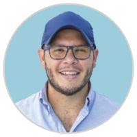 Hombre sonriente con gorra de béisbol azul, gafas y camisa azul claro abotonada, sobre un fondo circular azul claro.