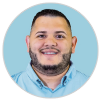 Hombre sonriente con pelo corto oscuro y barba, vestido con camisa de cuello azul claro, delante de un fondo azul claro.
