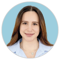 Una mujer de pelo largo y castaño sonríe a la cámara. Lleva una camisa de cuello azul claro y un collar. El fondo es de color azul claro sólido dentro de un marco circular.